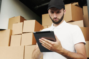 A delivery man reading a tablet standing behind a truckload of boxes.
