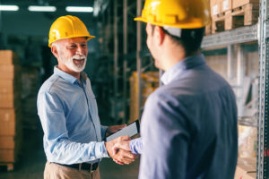 Businessmen wearing hardhats shaking hands in a warehouse.