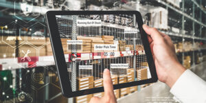 Image of a man's hands holding an electronic tablet in front of a warehouse shelf with inventory status displayed on it.