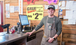 Smiling young man wearing apron stands next to cash register